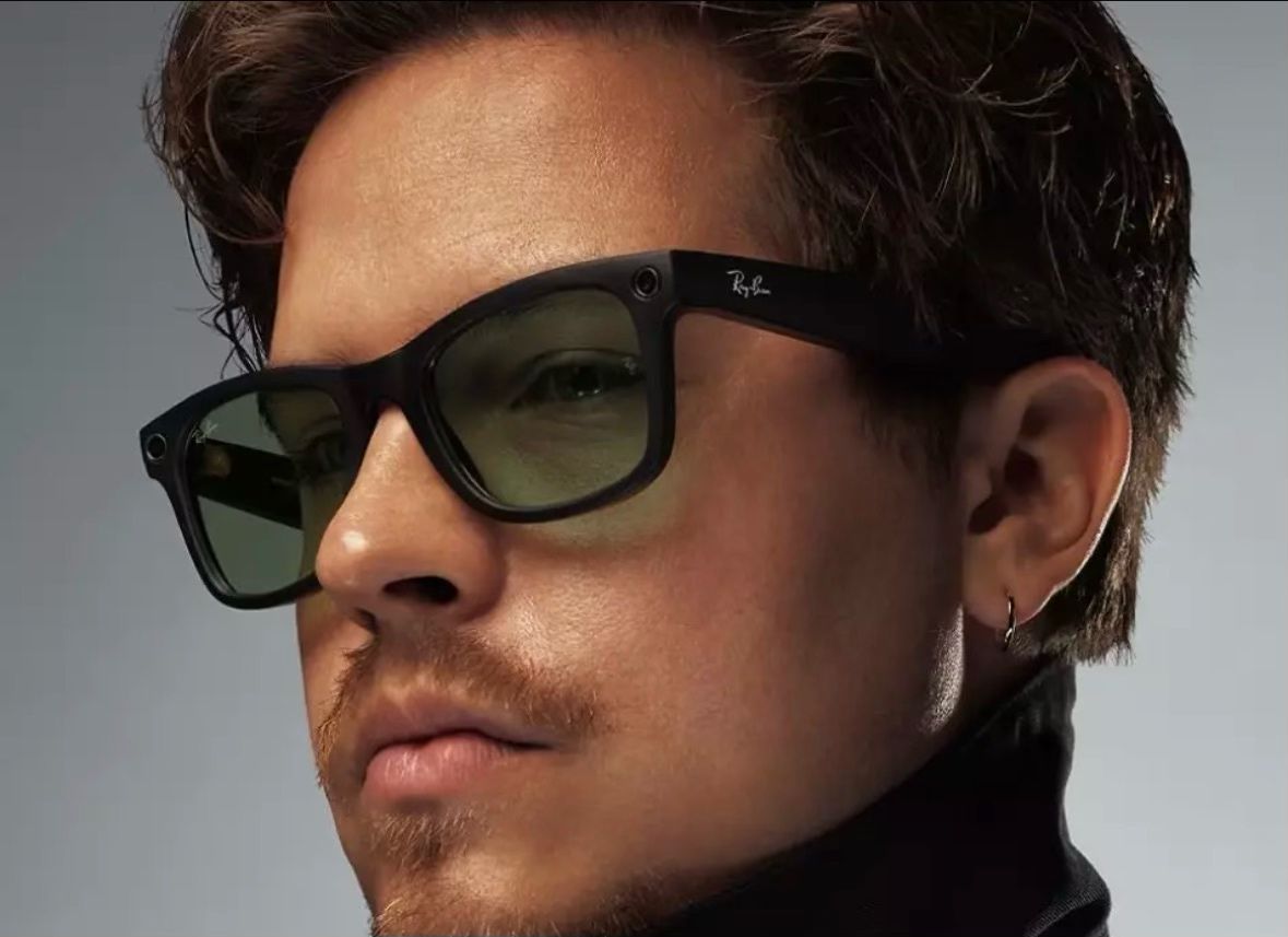 A close-up product shot of a man wearing black Ray-Ban Meta smart glasses, with the camera visible in the corner of the frame